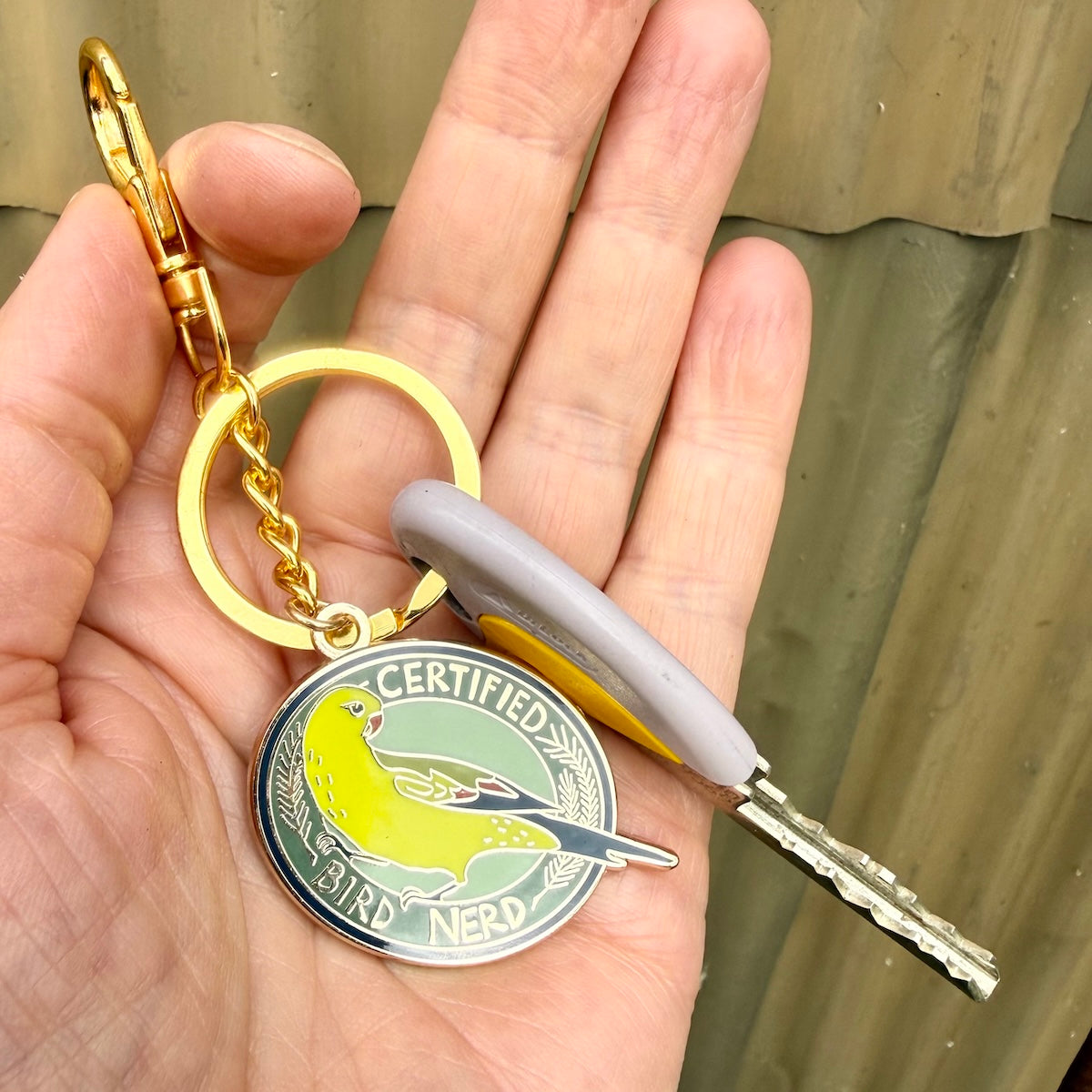 A gold-tone keychain with a round enamel bird charm reading 'CERTIFIED BIRD NERD' is held in a person's hand. The keychain includes a gold split ring, gold clasp, and is attached to a gray and yellow car key.