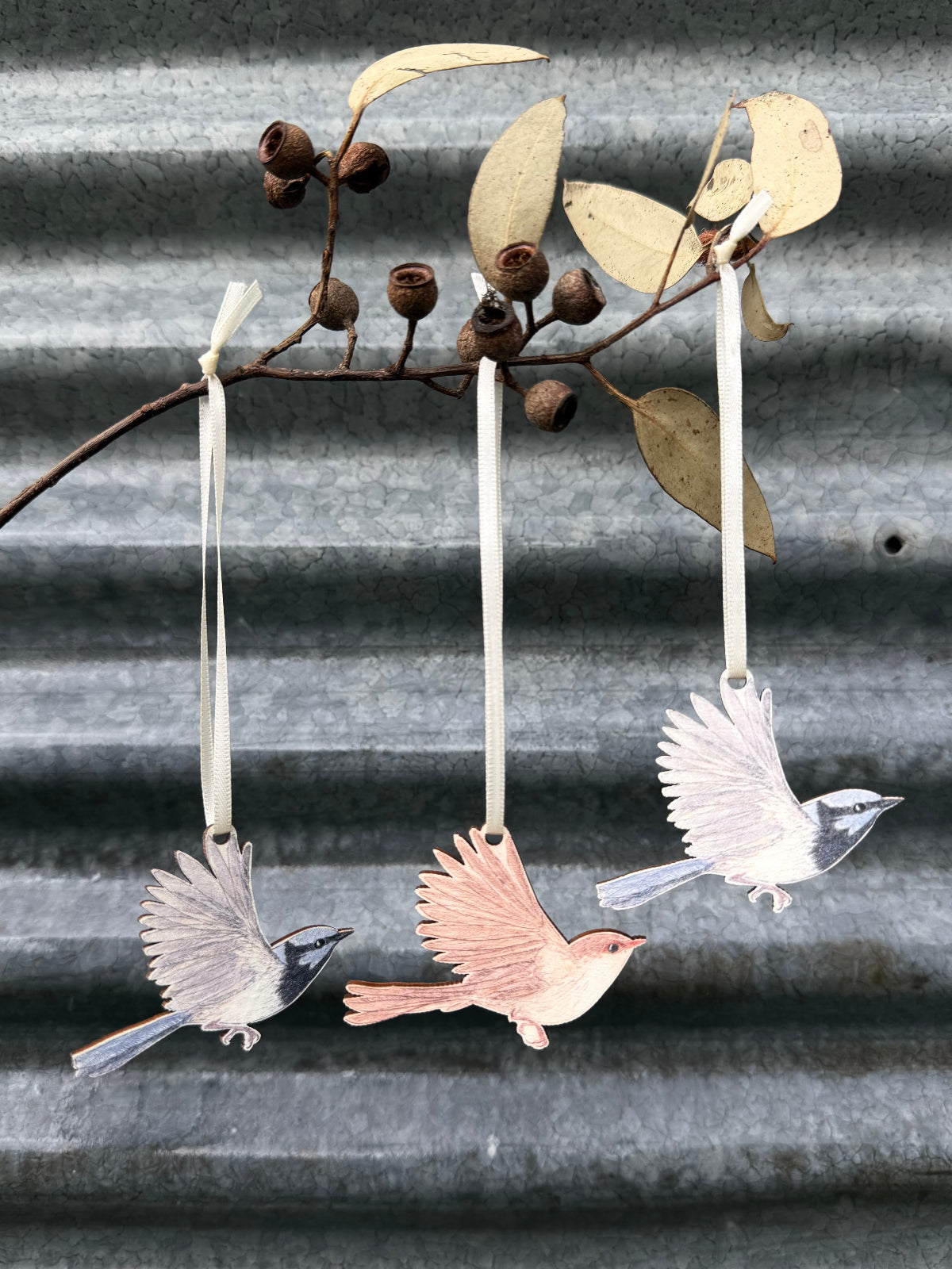 Three superb fairywren ornaments suspended from a dried eucalyptus branch against a corrugated metal wall.
