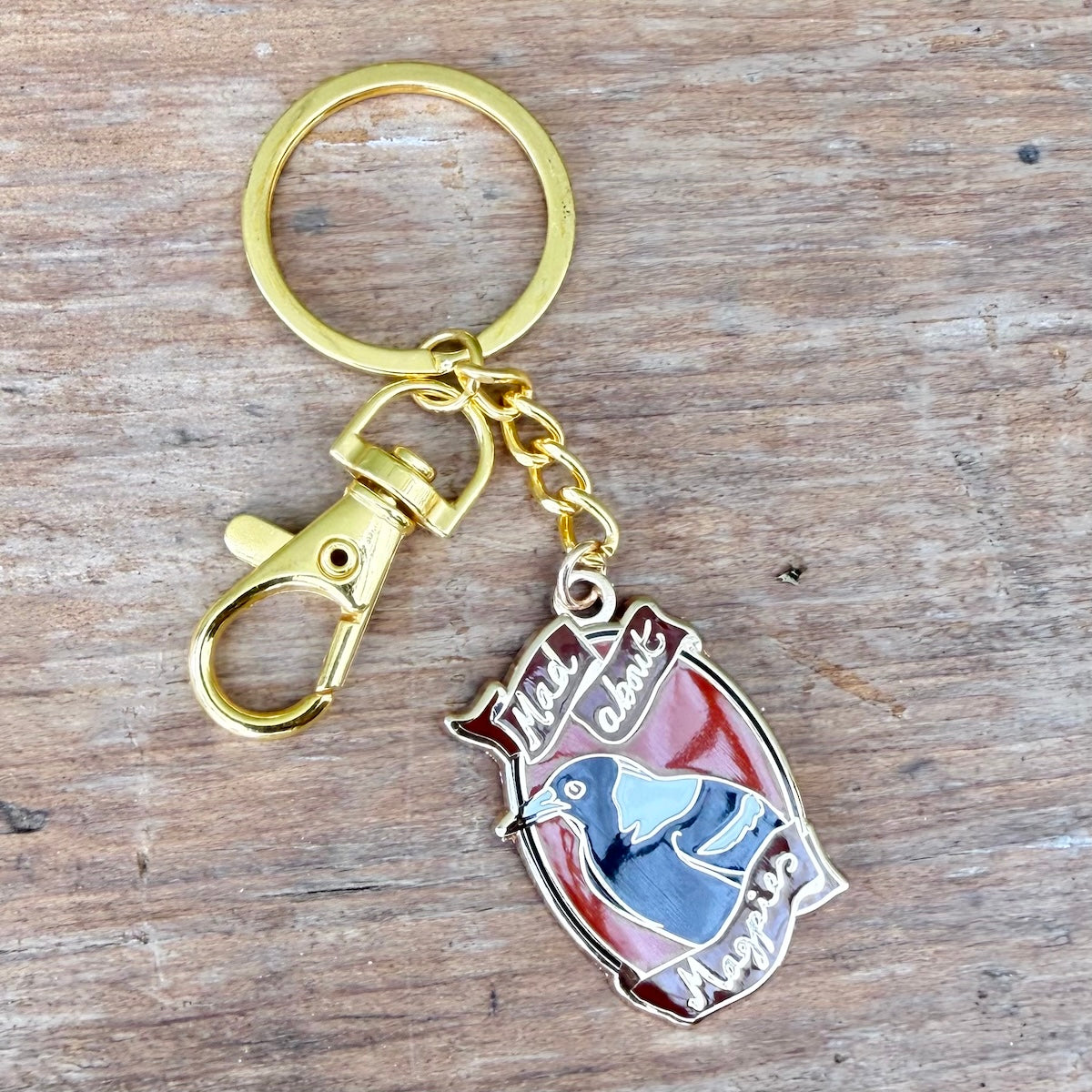 A gold-tone metal keyring with a lobster clasp and a shield-shaped charm featuring a magpie, displayed on a rustic wooden surface.