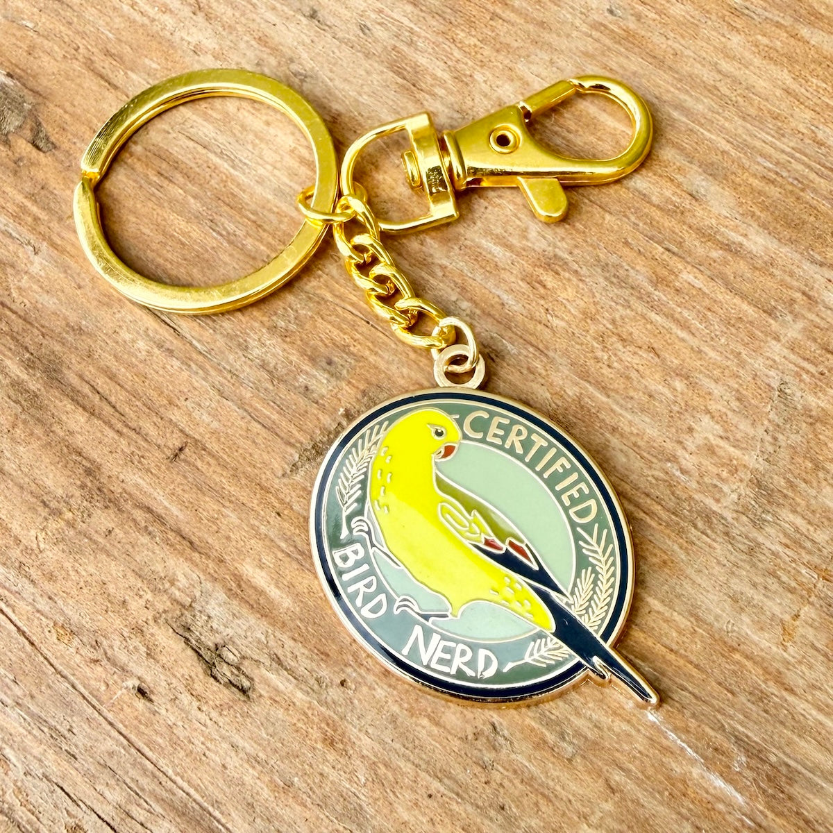 A gold-tone metal keychain with a lobster clasp and split ring, featuring a round enamel pin of a yellow bird on a light green background with the words 'CERTIFIED BIRD NERD' on a rustic wooden surface.