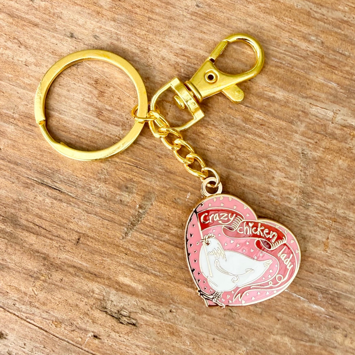 A gold-tone metal keychain with a lobster clasp and split ring, attached to a pink heart-shaped charm featuring a white chicken illustration and the words 'Crazy Chicken Lady'. The keychain is displayed on a rustic wooden surface.