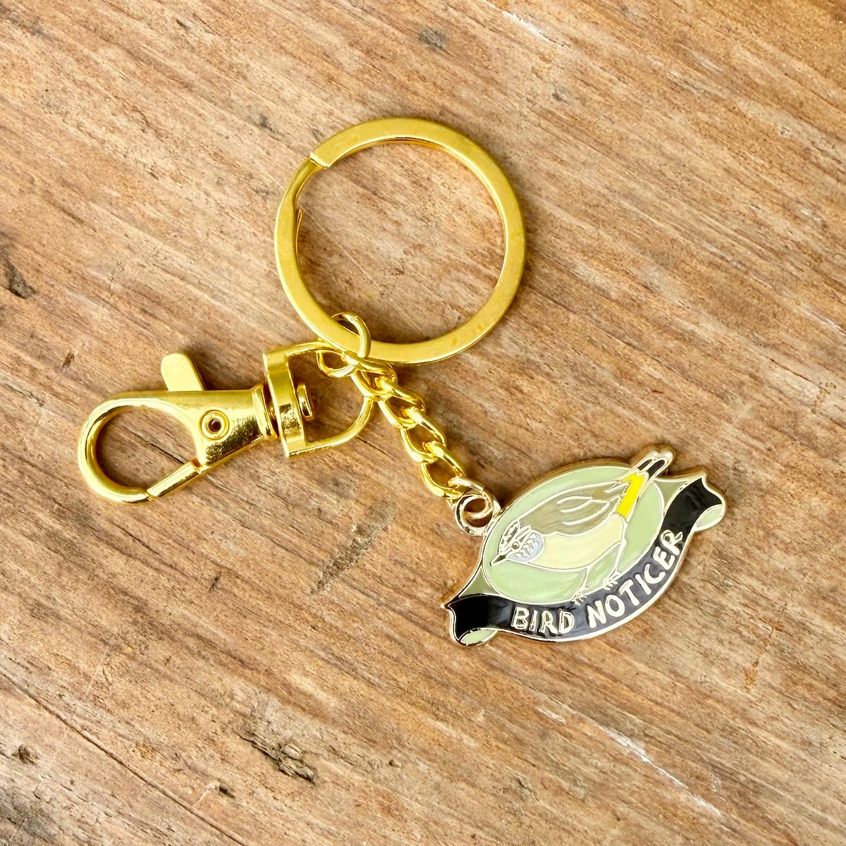 A gold-tone metal keychain with a lobster clasp and split ring, featuring an enamel bird charm labeled 'BIRD NOTICER' on a rustic wooden background.