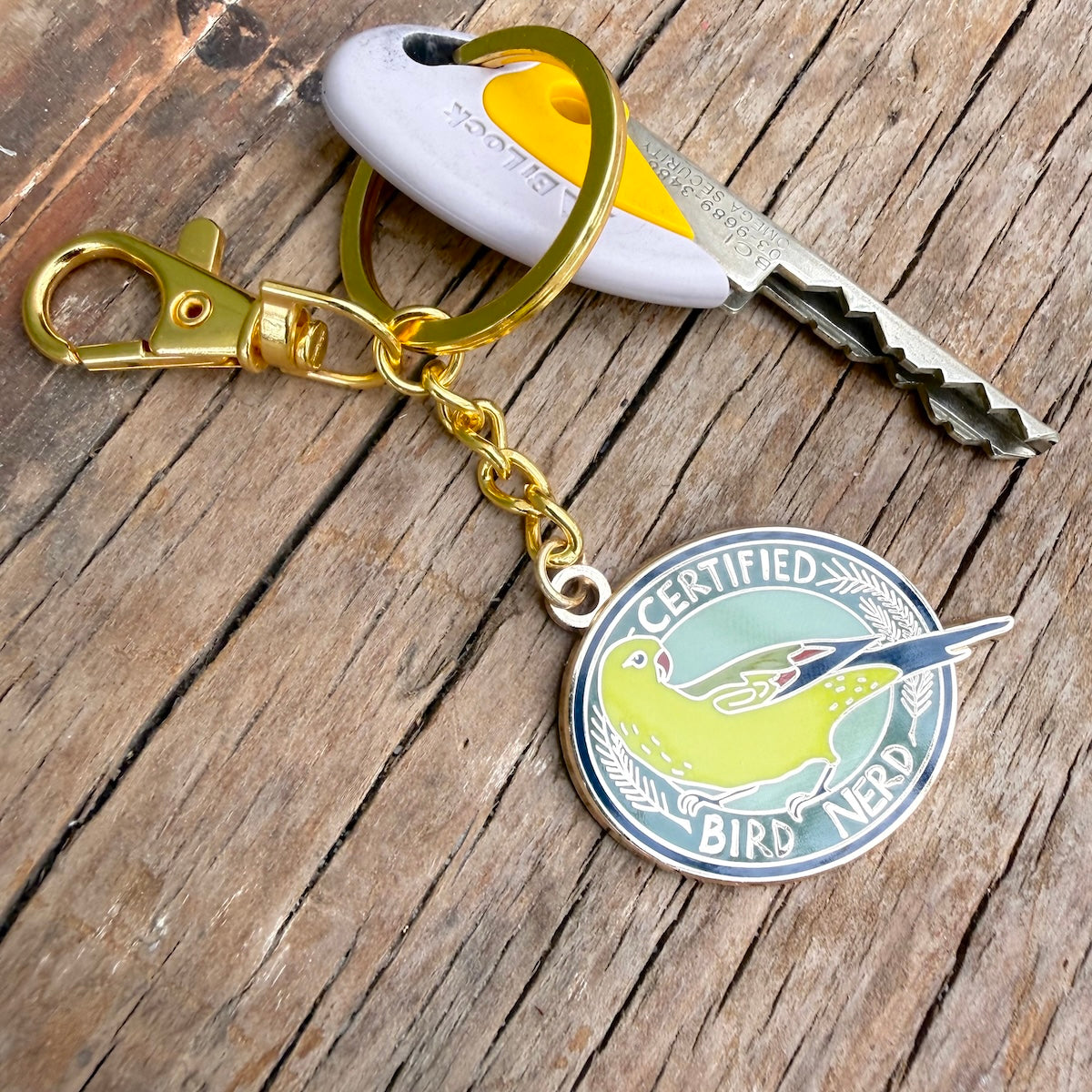 A gold-tone keychain with a lobster clasp, split ring, and round enamel bird-themed tag reading 'CERTIFIED BIRD NERD,' shown with keys on a rustic wooden surface.
