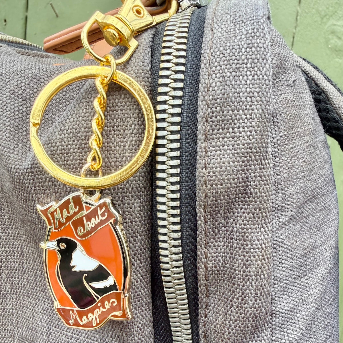 A close-up of a grey fabric zippered bag with a gold-tone keyring attached. The keyring features a circular ring, a chain, and an enamel charm depicting a magpie with the words 'Mad About Magpies' in gold script on an orange background.