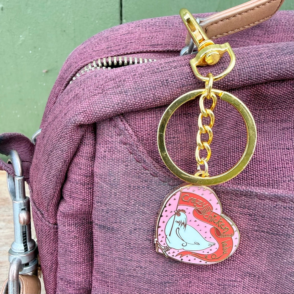 A close-up of a pink enamel keychain charm with a light blue seal and red banner reading 'Crazy Chicken Lady' attached to a gold-tone ring and clasp, hanging from a mauve fabric bag.