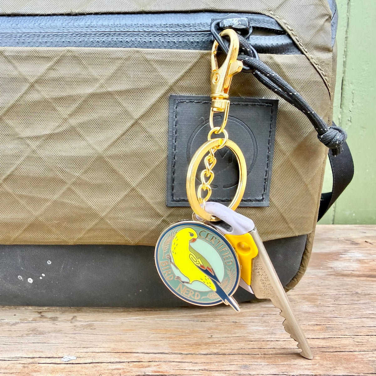 A close-up of a tan quilted bag with a black strap and a gold-tone keyring accessory hanging from it. The keychain includes a round enamel pin featuring a yellow bird and the words 'Certified Bird Nerd' and a key attached.