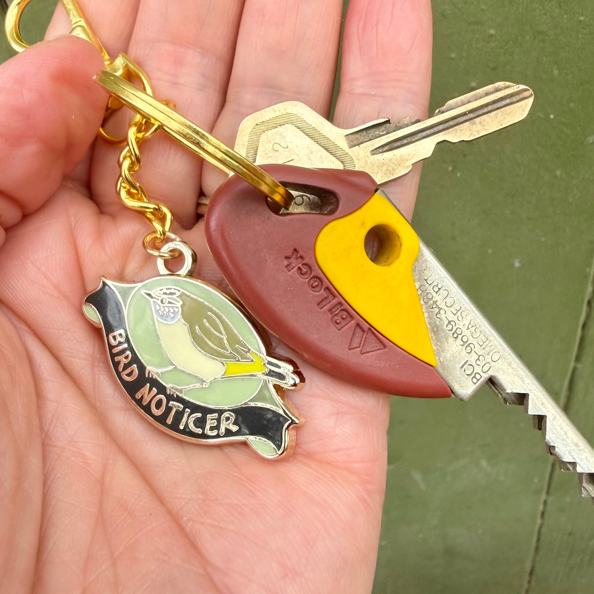A hand holding a gold-tone keyring with two keys. One key has a brown and yellow plastic key fob, and the other is a bird-shaped keychain labeled 'BIRD NOTICER' with a small bird illustration.