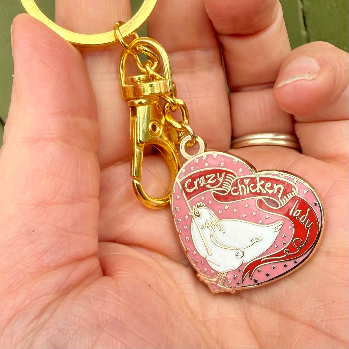 A close-up of a hand holding a gold-tone keychain with a heart-shaped charm. The charm has a pink background with a white chicken illustration and the words 'Crazy Chicken Lady' in red script.