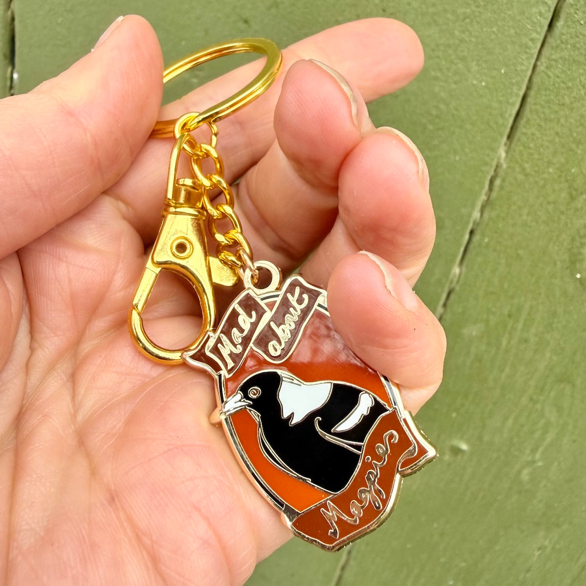 A hand holding a small enamel keyring featuring a black and white bird on an orange shield with the words 'Mad About Magpies' and a gold clasp, against a green painted wooden background.