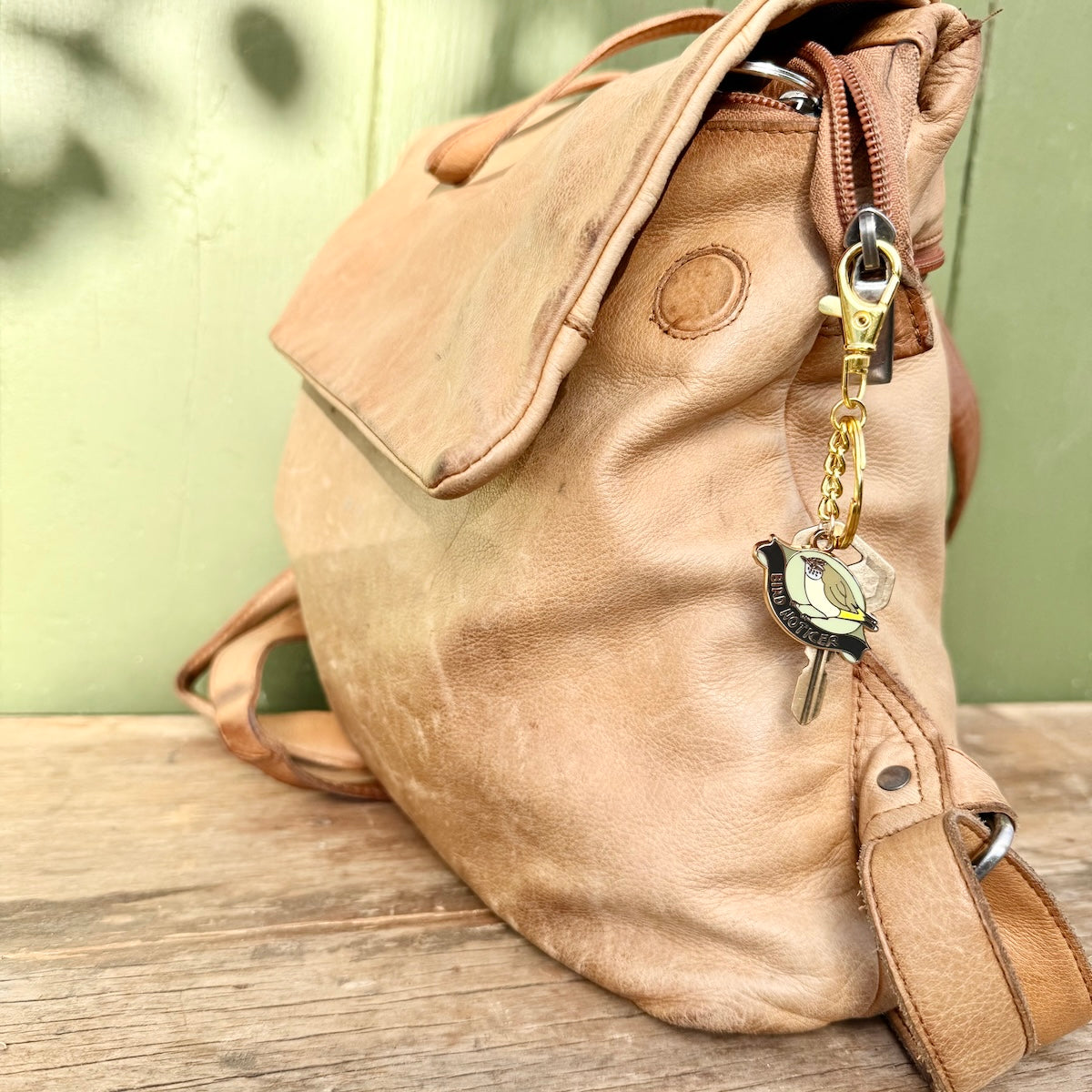 A light tan leather shoulder bag with a flap closure, shown resting on a rustic wooden surface against a pale green wall. The bag features a gold-tone keychain with a bird charm.