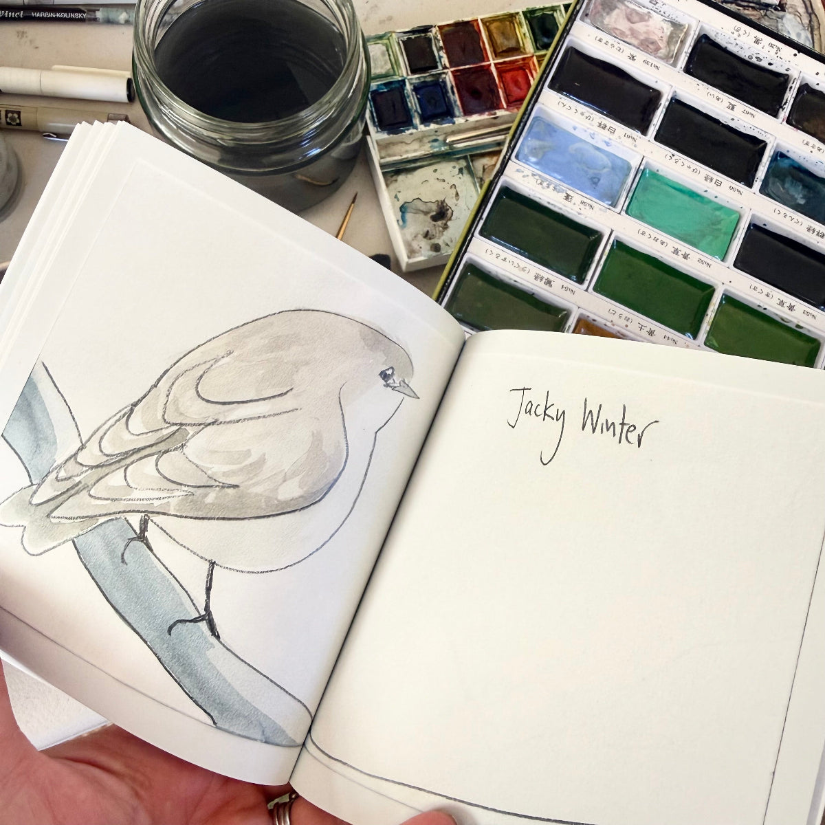 Open sketchbook with watercolor painting of a bird and art supplies on a table.