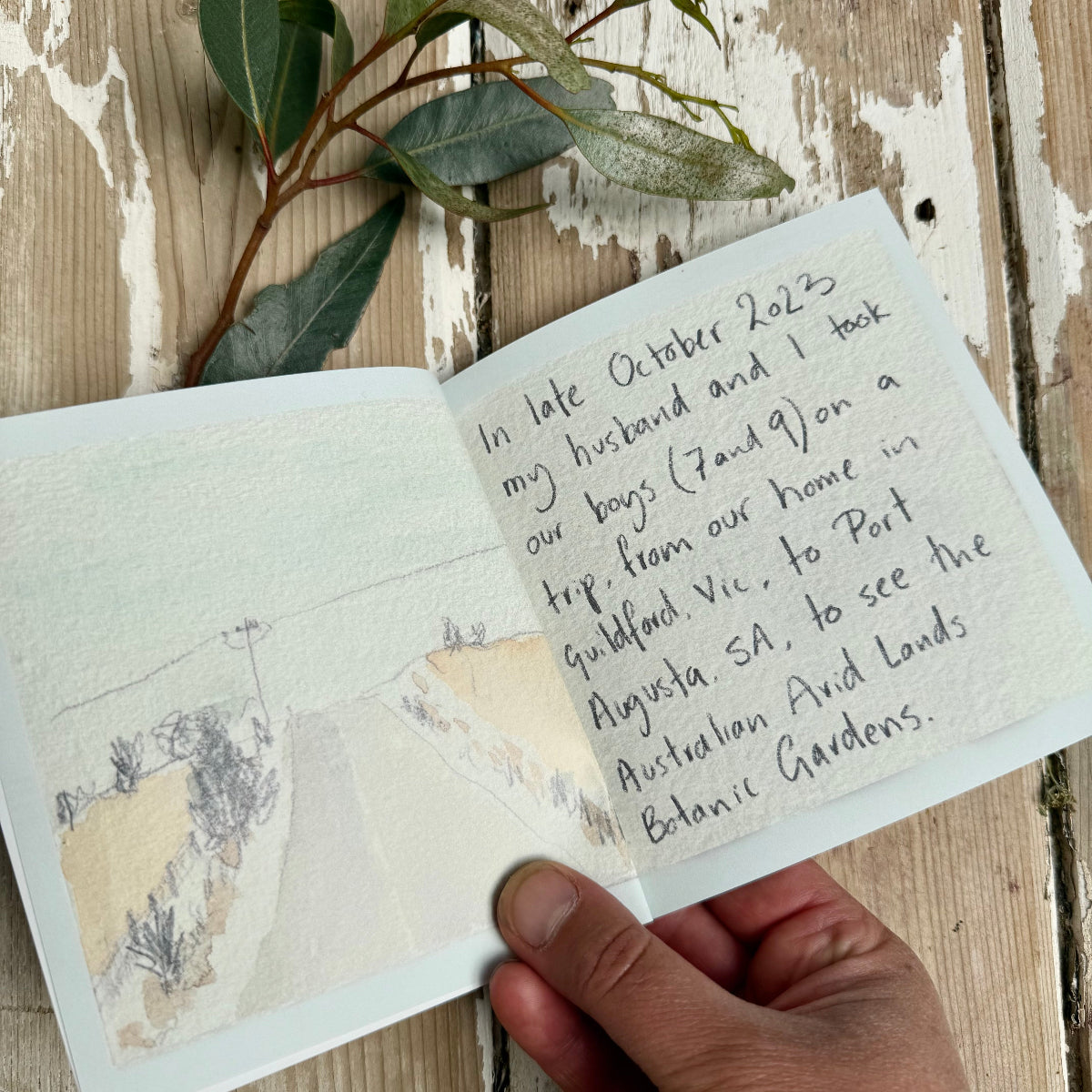 Handwritten note on a small book with a scenic illustration, held on a wooden surface with green leaves.