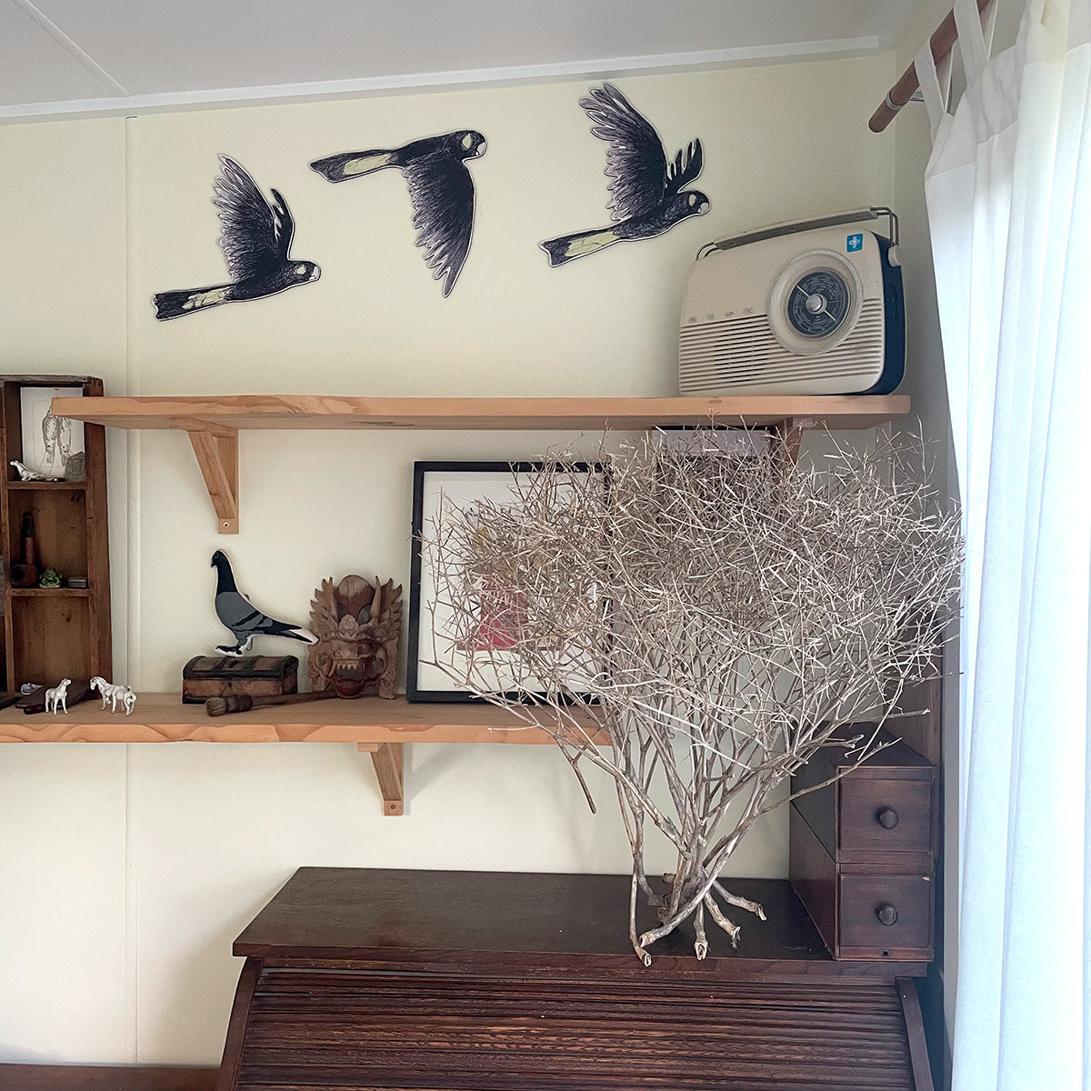 A styled interior corner with cream walls, wooden floating shelves, black bird wall decals, a vintage radio, framed art, and a large dried branch arrangement on a dark wooden cabinet.